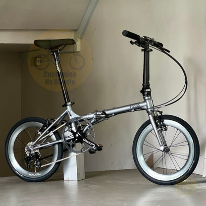 A silver folding bicycle with black components, including the saddle, handlebars, and tires, displayed indoors on a concrete floor against a beige wall.