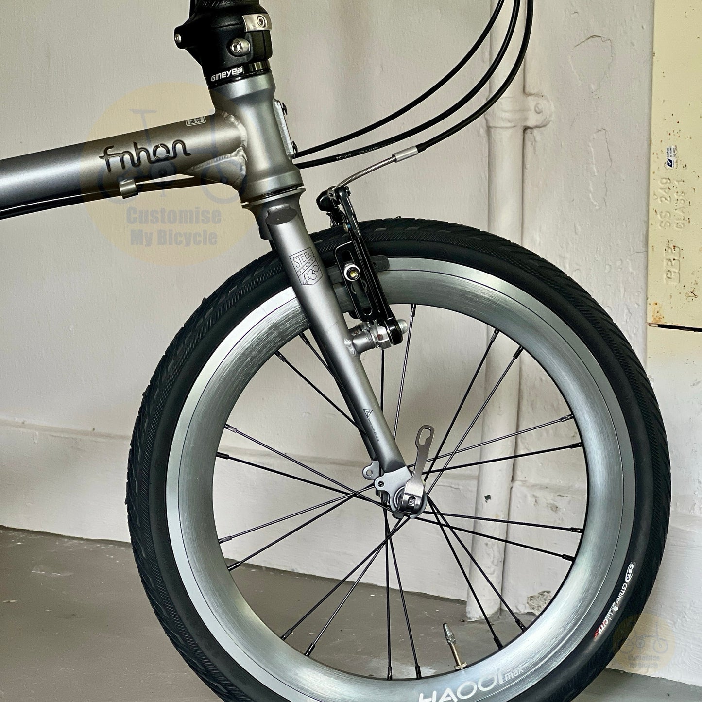 Close-up of a folding bicycle wheel and front fork, showing a silver aluminum rim, black tire, and metallic components against a white wall.