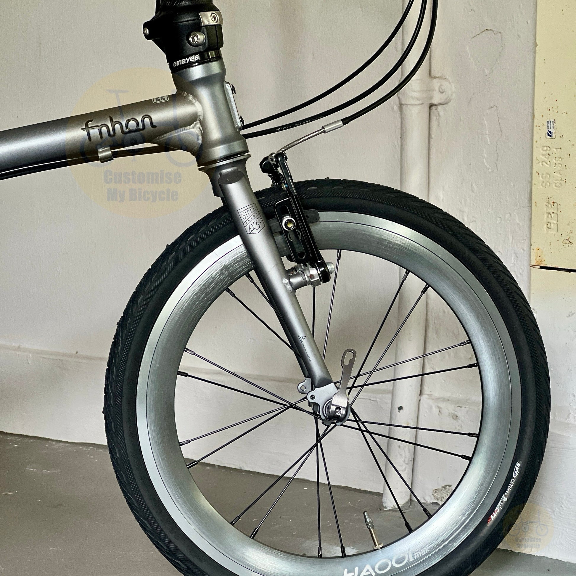 Close-up of a folding bicycle wheel and front fork, showing a silver aluminum rim, black tire, and metallic components against a white wall.