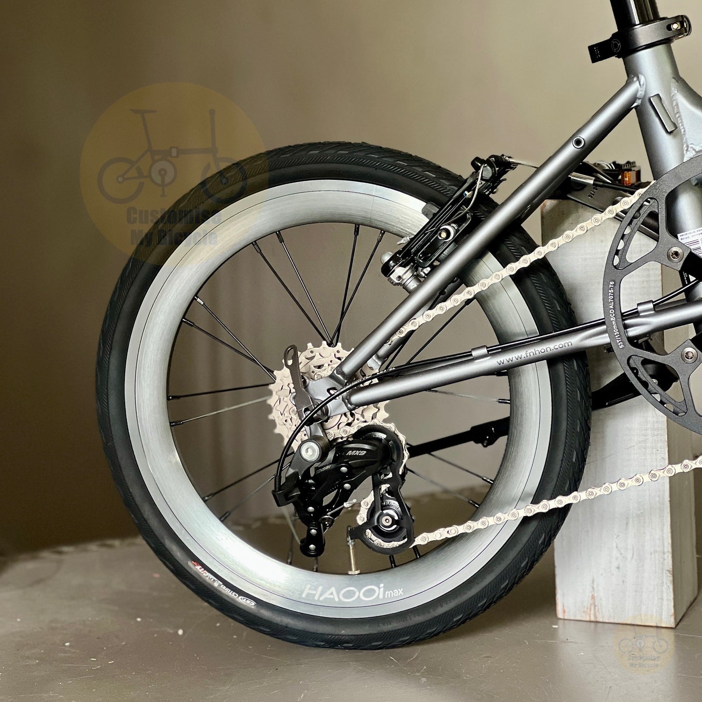 Close-up of a bicycle rear wheel and frame, showing a silver-gray metal frame, black tire, silver rim, rear derailleur, chainring, and chain. The bike is photographed indoors against a neutral beige wall and floor, resting against a white wooden block.