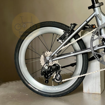 Close-up of a bicycle rear wheel and frame, showing a silver-gray metal frame, black tire, silver rim, rear derailleur, chainring, and chain. The bike is photographed indoors against a neutral beige wall and floor, resting against a white wooden block.