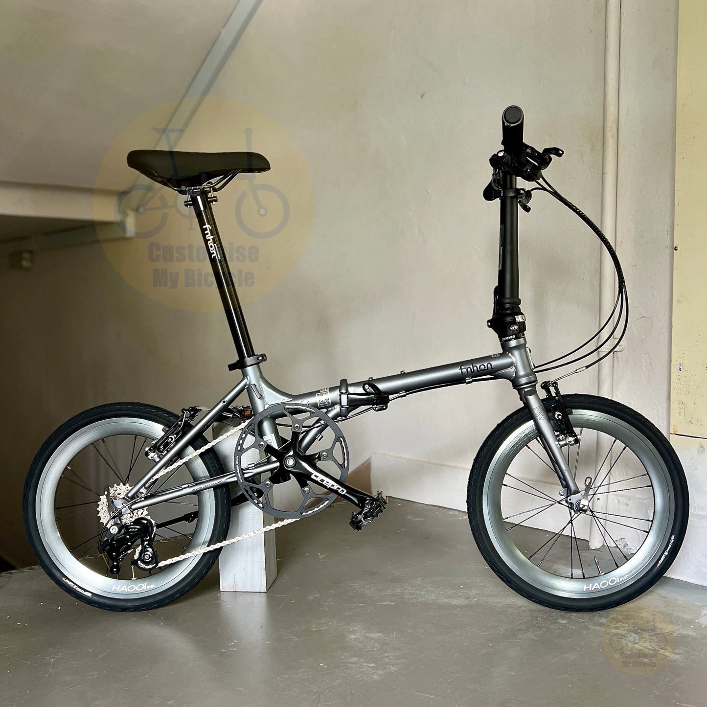 A silver folding bicycle with black tires and components, photographed indoors against a beige wall and concrete floor. The brand name 'BONSAI' is visible on the frame.
