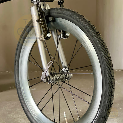 Close-up of a bicycle front wheel with a silver rim, black tire, silver spokes, and silver fork against an indoor neutral background.