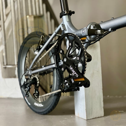 Close-up of a bicycle crankset and rear wheel with a matte gray frame, black chainring, and visible 'UCITO' branding, photographed indoors against a neutral wall and floor.