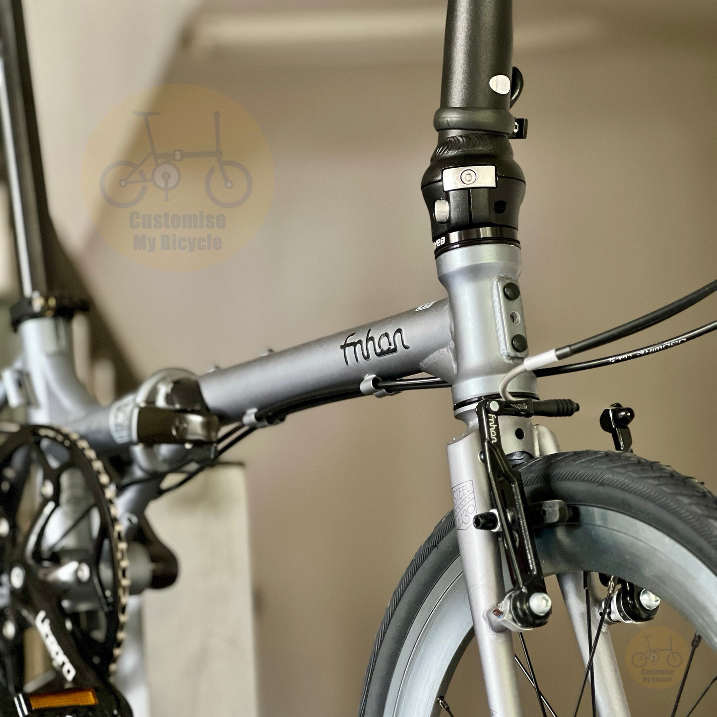 Close-up of a folding bicycle frame in metallic silver with black components, including the fork, brake levers, and chainring area. The frame has the brand name 'fihan' written on it.