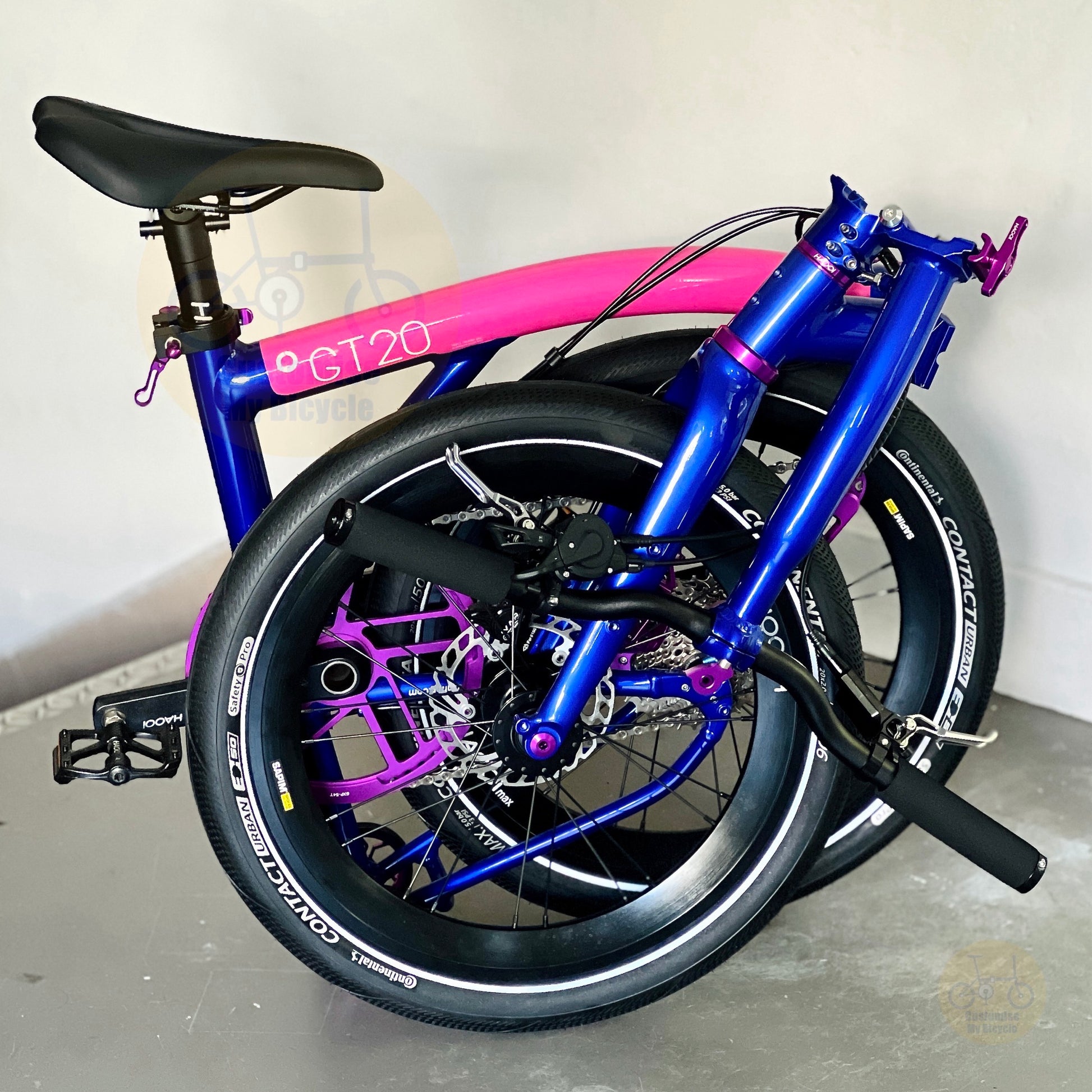 A compact folding bicycle with a vibrant metallic blue frame and a bright pink top tube, featuring purple accents on the fork and seat post, photographed on a concrete floor against a plain wall.