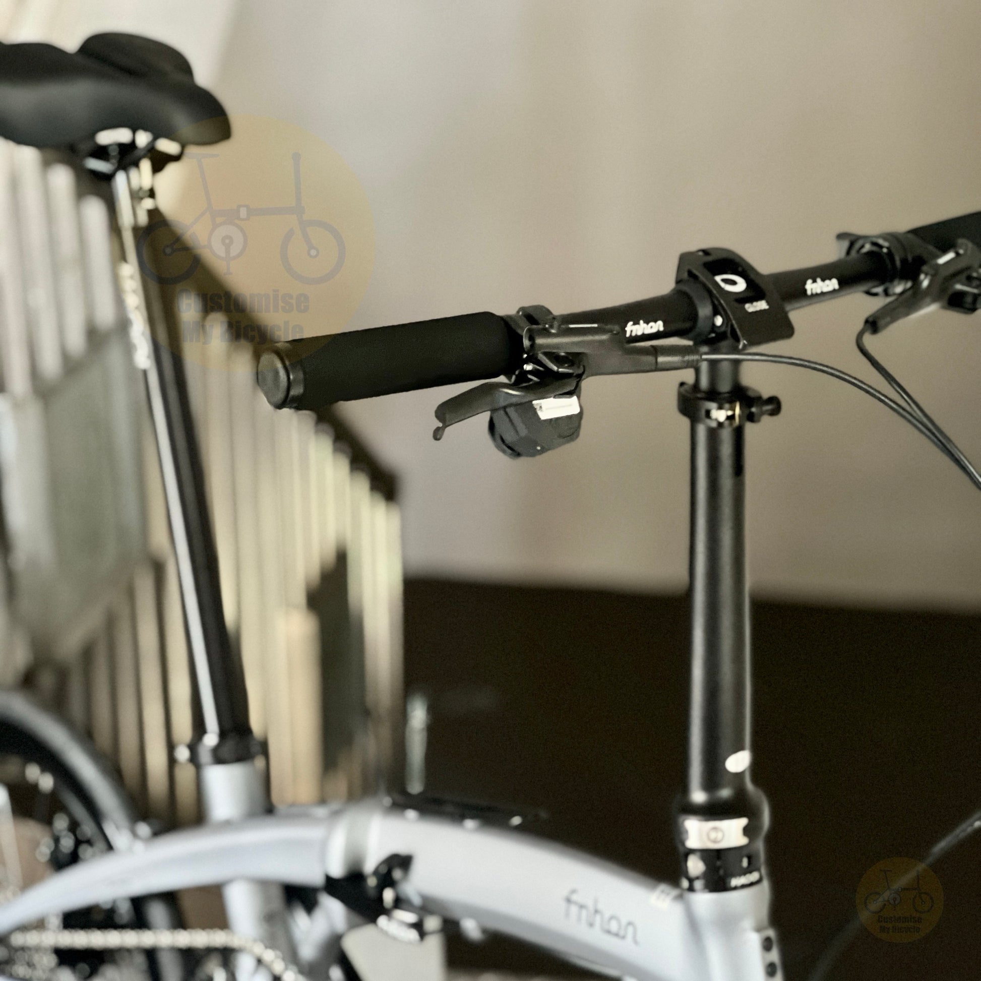 Close-up of a folding bicycle handlebar area with black grips, black handlebar clamp, and a light gray frame. The brand name 'fnhon' is visible on the frame and handlebar.