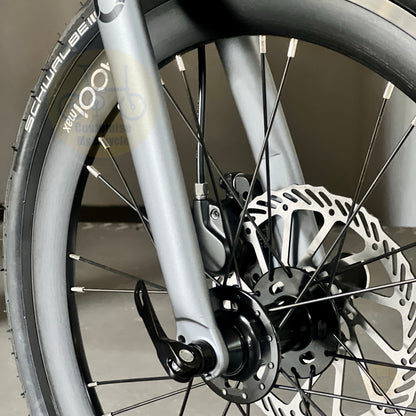 Close-up of a bicycle wheel and front fork showing a silver disc brake rotor, black spokes, and part of a black tire with white branding text.